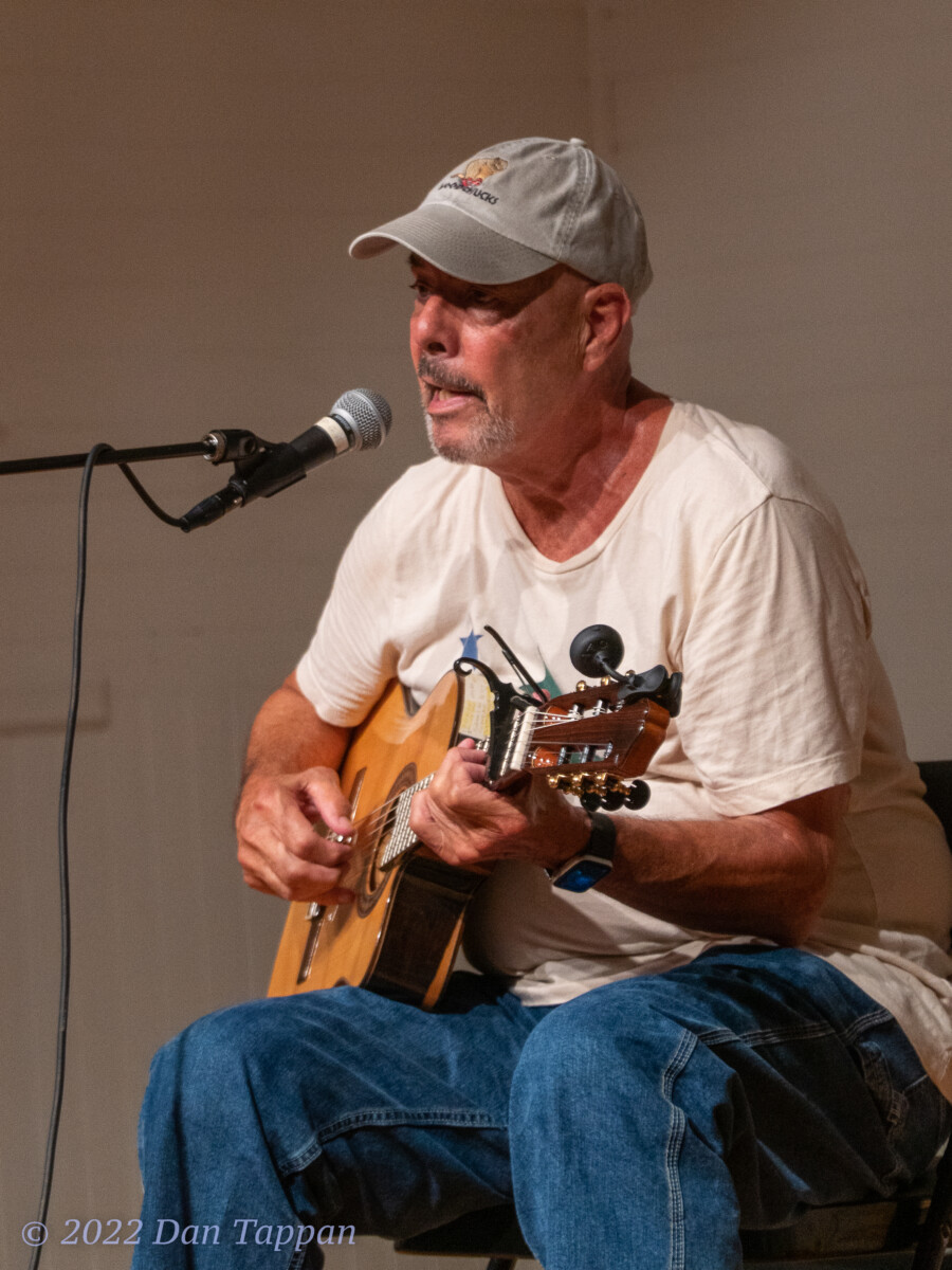 Musician John Schindler, wearing a t-shirt and baseball cap and holding a guitar, performing at the PCA Open Mike